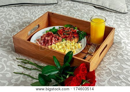 Breakfast in bed on wooden tray with bunch of red roses. Scrumbled eggs, fried bacon, beans on toast and salad, with orange juice.