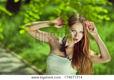 Close-up horizontal portrait of sensuality young woman with your hands near the headlong hair a red lips a thoughtful gaze at the open air in the middle of green blurred leaves at sunny spring day