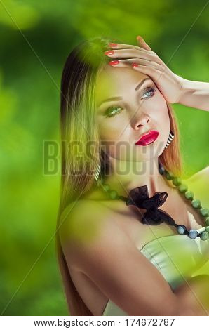 Close-up portrait of sensuality young woman with your hands near the headlong hair a red lips a thoughtful gaze at the open air in the middle of green blurred leaves at sunny spring day