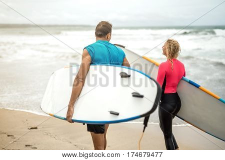 Athletic couple of surfers walks with surfboards on the beach on the background of the ocean waves and the sky. Girl wears a black-pink swimsuit. Guy wears a cyan T-shirt and black shorts.