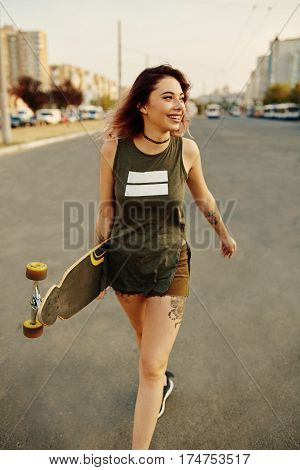 Beautiful young tattooed woman with her longboard on the road in the city in sunny weather