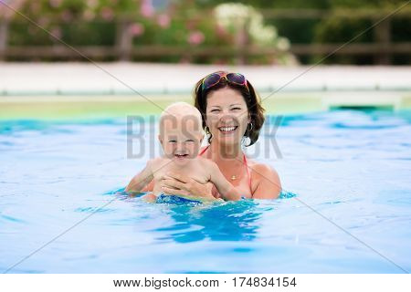 Happy young mother playing with her baby in outdoor swimming pool on hot summer day. Kids learn to swim during family vacation. Children relaxing in tropical resort. Mom and child play in water.