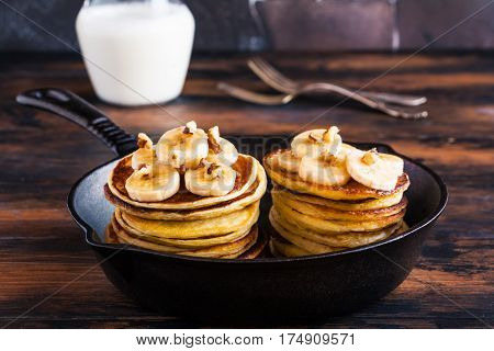 Stack of homemade pancakes with banana maple syrup and walnuts in black cast iron skillet. Vintage forks glass jar with milk wooden rustic table.