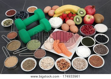 Body building dumbbell weights with food of supplement powders, high protein lean meat and salmon, fruit, dairy, nuts, seeds and grains over slate background.