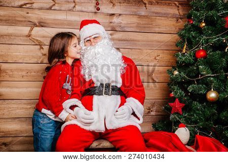 Santa Claus And Little Girl. Girl Telling Wish In Santa Clauss Ear In Front Of Christmas Tree
