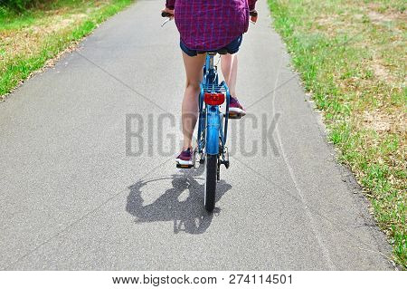 Young Woman Is Riding A Blue Bicycle In A Purple Shirt. View From The Back. Saddle And Wheel