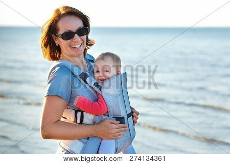 Baby And Mother On Sea At Summer Day. Child In A Carrier Backpack
