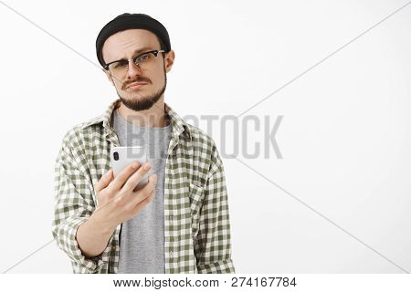 Indifferent Male Young Snob In Glasses And Beanie With Beard Holding Smartphone Smirking And Staring