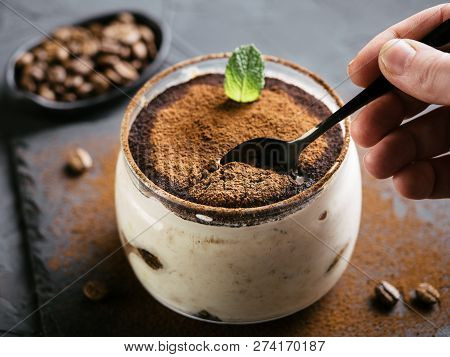 High Angle View Of Tiramisu In Glass Cup On Stone Plate Over Black Concrete Background, Hand With Te