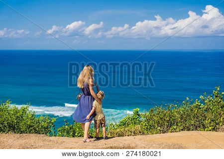 Mom And Son Travelers On A Cliff Above The Beach. Empty Paradise Beach, Blue Sea Waves In Bali Islan