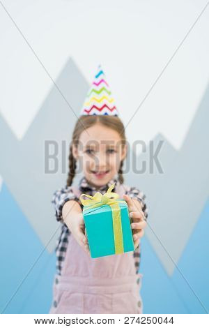 Positive Cute Girl In Party Hat Standing Against Wall With Mountains Drawing And Giving Blue Gift Bo