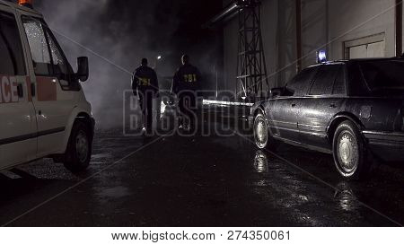 Fbi Agents Work At The Scene At Night, Police Car With Lights And Ambulance Background. Back View On