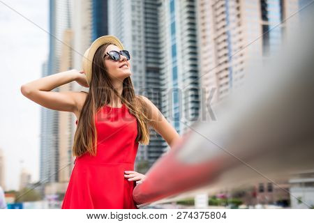 Portrait Of Young Woman Wearing In Red Dress And Sunglasses, Straw Hat In Front Of Skycrapers In Mod