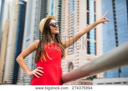 Young Pretty Woman In Red Dress And Straw Hat Pointed With Hands On Downtown Skycrapers In Modern Ci