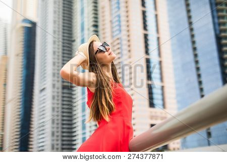 Young Pretty Woman Holding Straw Hat Wearing In Red Dress And Sunglasses In Front Of Skycrapers.