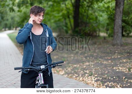 Attractive Young Caucasian Man With Dark Hair Bicycling In The Park. Touches The White Earphones. Ou