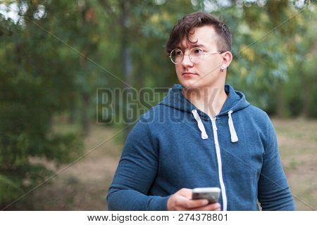 Attractive Young Caucasian Man With Dark Curly Hair, Golden Round Glasses And Earring Holds The Mobi