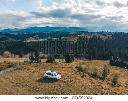 Aerial View Of Man In Red Coat Near White Suv Car At Hill With Beautiful Mountains View. Road Trip.