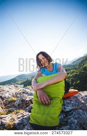 A Woman Is Sitting In A Sleeping Bag At Sunset. Camping On Top Of A Mountain. A Smiling Girl Rests A