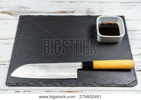 Black Stone Plate Board On A White Rusty Background With A Sushi And Sashimi Knive On It.