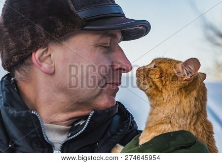 A Middle-aged Man In A Brown Hat And A Red Cat Look At Each Other In Winter