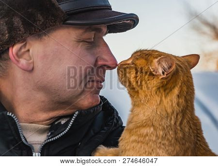 A Middle-aged Man In A Brown Hat And A Red Cat Look At Each Other In Winter