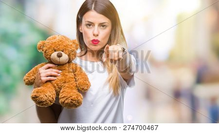 Young beautiful woman holding teddy bear plush over isolated background pointing with finger to the camera and to you, hand sign, positive and confident gesture from the front