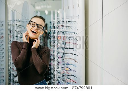 Optics. Close-up Of A Young Beautiful Girl Wearing Glasses, Smiling Near The Stand. Ptical Store.