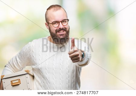 Young hipster man wearing glasses, moving holding moving box over isolated background happy with big smile doing ok sign, thumb up with fingers, excellent sign