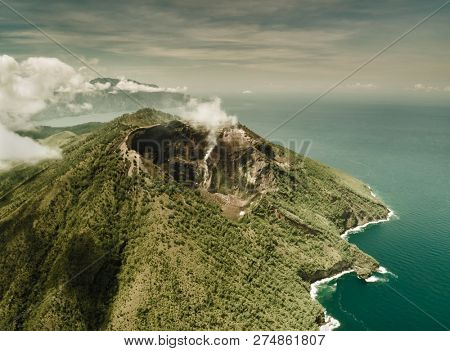 Overview of active Indonesia volcano. Aerial drone shot. Overwhelming scenery of Indonesian nature. Greens covered land surrounded by the ocean. Beauty of untouched Asian environment. Exotic tourism.