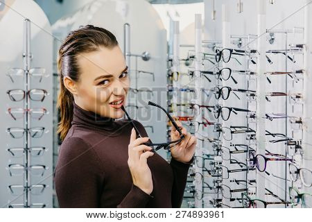 Optics. Close-up Of A Young Beautiful Girl Wearing Glasses, Smiling Near The Stand. Ptical Store.