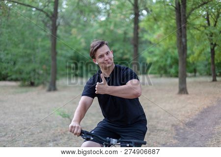 Young Handsome Caucasian Man Thumbs Up Bicycling At The Park. White Earphones, Dark Casual Wear. Out