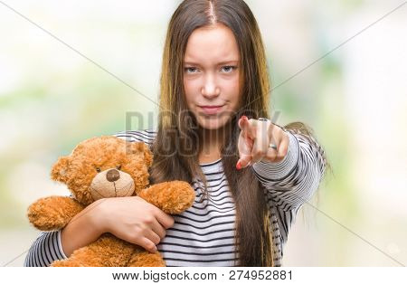Young caucasian woman holding teddy bear over isolated background pointing with finger to the camera and to you, hand sign, positive and confident gesture from the front