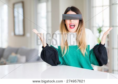 Young beautiful woman blindfold eyes with black censor band at home celebrating crazy and amazed for success with arms raised and open eyes screaming excited. Winner concept