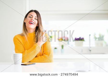 Young beautiful woman drinking a cup of coffee at home cheerful with a smile of face pointing with hand and finger up to the side with happy and natural expression on face