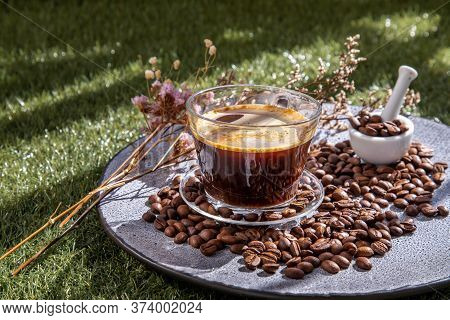 Coffee Style, Black Coffee In Glass Cup With Coffee Beans Roating And Dried Flower On Ceramic Plate 