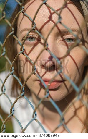 Close-up Face Of A Young Girl Looking Through A Metal Fence.
