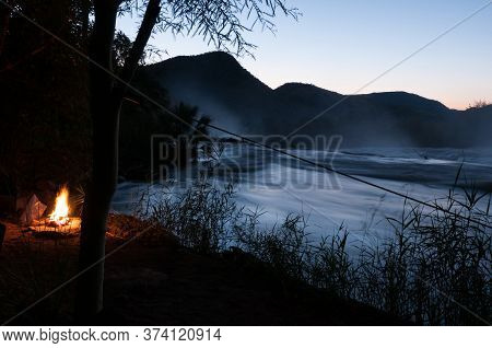 A Campfire At Sunset Next To The Kunene River At The Top Of The Epupa Waterfalls