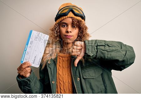 Young african american tourist woman wearing ski goggles holding plane boarding pass tickets with angry face, negative sign showing dislike with thumbs down, rejection concept