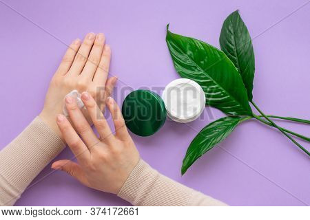 Girls Hand Is Nearby Jar With White Soft Hand And Body Cream On Purple Background With Large Green L
