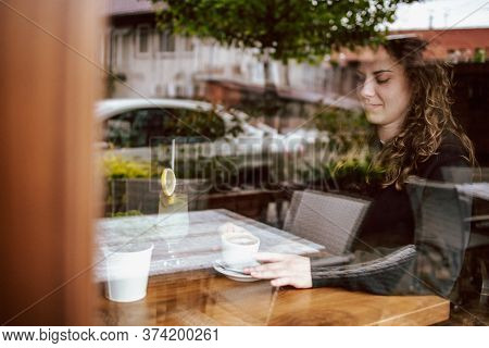 Cute Brunette, Curly Girl Sitting In A Cafe, Drinking Coffee And Juice. View Though The Wondow