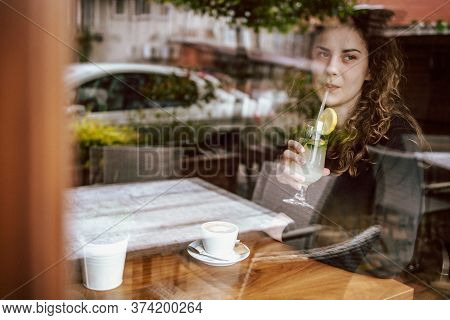 Cute Brunette, Curly Girl Sitting In A Cafe, Drinking Coffee And Juice. View Though The Wondow