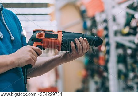 A Man Holds An Electric Drill In His Hand For Drilling. A Buyer In A Hardware Store Selects A Produc