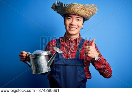 Young handsome chinese farmer man wearing apron and straw hat holding watering can happy with big smile doing ok sign, thumb up with fingers, excellent sign