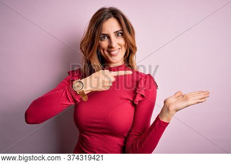 Young beautiful brunette woman wearing casual sweater stnding over pink background amazed and smiling to the camera while presenting with hand and pointing with finger.