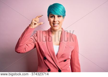 Young beautiful businesswoman with blue fashion hair wearing jacket over pink background smiling and confident gesturing with hand doing small size sign with fingers looking and the camera. Measure