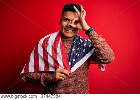 Young handsome latin patriotic man wearing united states of america falg with happy face smiling doing ok sign with hand on eye looking through fingers