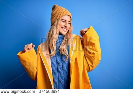 Young beautiful blonde woman wearing raincoat for rainy weather over blue background very happy and excited doing winner gesture with arms raised, smiling and screaming for success. Celebratio.