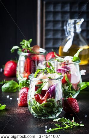 Summer Salad With Arugula, Soft Cheese, Red Strawberry And Prosciutto In Glass Jars On Black Table, 