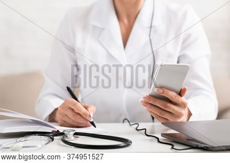 Doctor Work Remotely Online. Woman Doctor In White Coat Makes Notes In Notebook, Near Are Stethoscop
